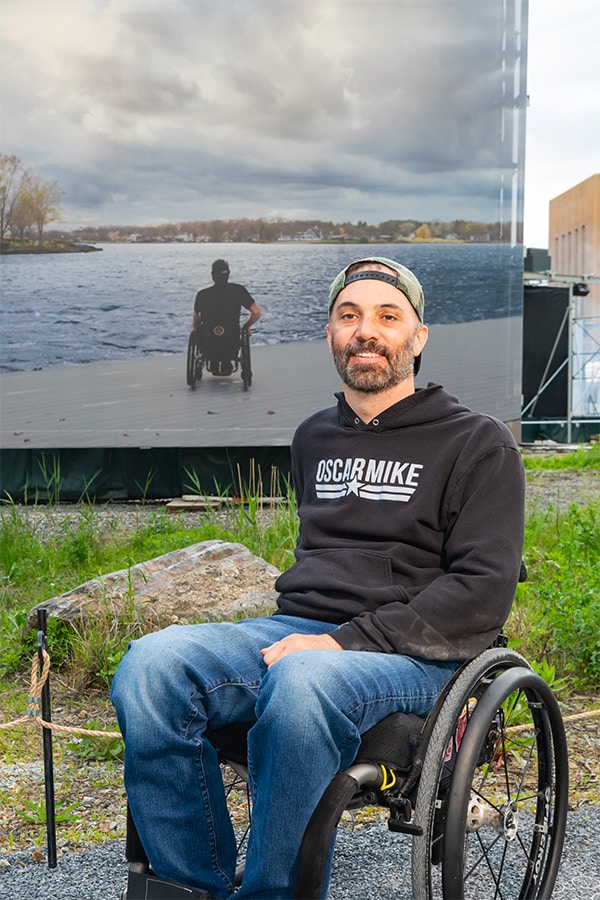 Retrato de um homem em cadeira de rodas vestindo um moletom “Oscar Mike”, sentado em frente a uma fotografia sua olhando para um lago, exibida ao ar livre na instalação “Path of Liberty” em Nova York.