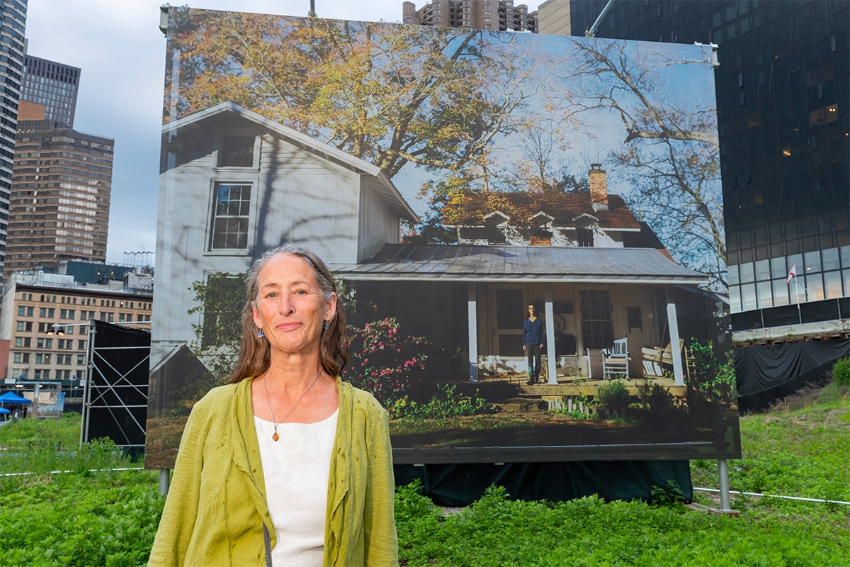 Retrato de uma mulher em frente a uma grande imagem de uma casa histórica de madeira com uma pessoa na varanda, parte da exposição “Path of Liberty”.